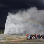 Old Faithful Geyser erupts with dark skies behind and a rainbow before, Upper Geyser Basin, Yellowstone National Park, Wyoming