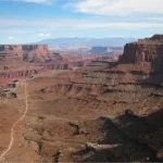 A road winds through a deep desert canyon with mountains and clouds beyond