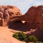 Rock rocks rise in lumpy domes around an arch while a hiker walks across slickrock