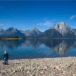 A hiker stands beside a large lake with reflections of the Teton Range, most notably Mount Moran on the right