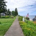 A sidewalk travels between a lawn and a garden with views out to a lake