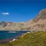 An alpine lake in a meadow bordered by mountains with a hiker looking on