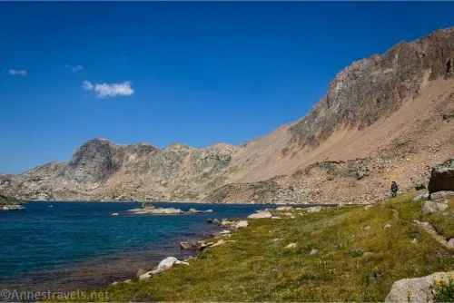 An alpine lake in a meadow bordered by mountains with a hiker looking on