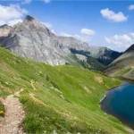 A trail on a green grassy hillside above a blue lake with rugged mountains and clouds in the distance