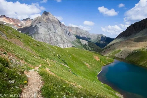 A trail on a green grassy hillside above a blue lake with rugged mountains and clouds in the distance