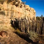 A corral made of upright branches and a metal burn barrel beside a yellow cliff face