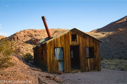 A wooden cabin with three windows and a missing door and a rusty chimney in front of desert hills