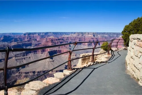 A railing along a paved path beside a wall with views to the Grand Canyon and blue sky beyond the railing