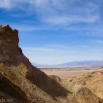 A rock formation rises from a yellow canyon with blue sky with clouds and distant mountains beyond