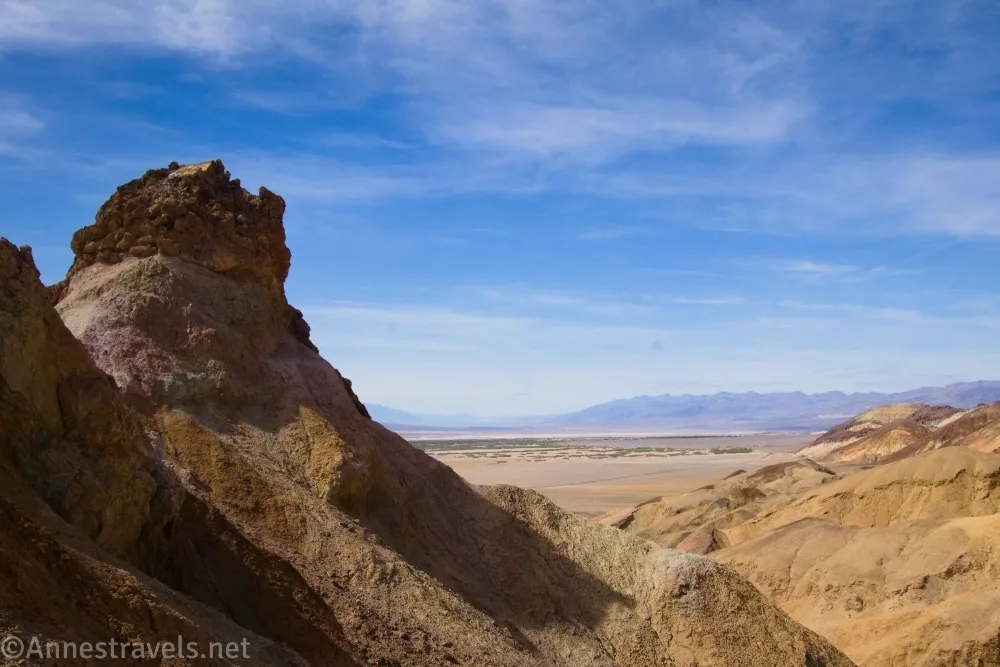 A rock formation rises from a yellow canyon with blue sky with clouds and distant mountains beyond