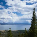 Trees rise in front of a view of a lake below clouds in a blue sky with mountains between the sky and the lake