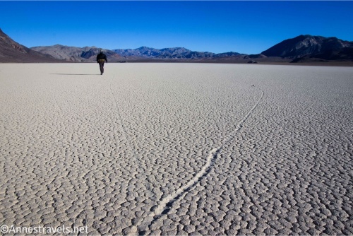 A hiker in a black jacket walking on a dry lakebed called the Racetrack Playa with the rock trails left from the Sailing Stones